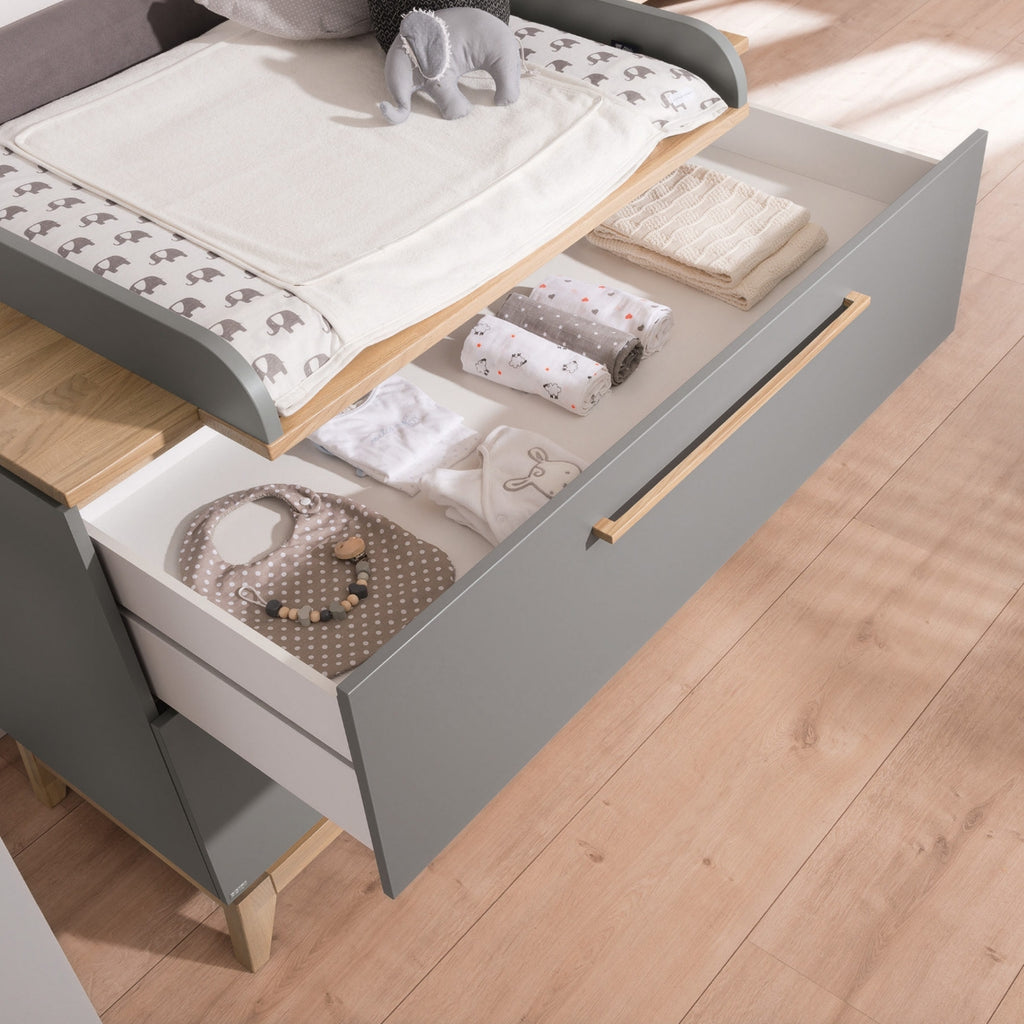 Offene, graue PAIDI Babyzimmer Sten – Wickelkommode mit Schubladen, in denen gefaltete Babykleidung und Spielzeug liegen, auf einem massiven Eichenboden.