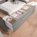 Offene, graue PAIDI Babyzimmer Sten – Wickelkommode mit Schubladen, in denen gefaltete Babykleidung und Spielzeug liegen, auf einem massiven Eichenboden.