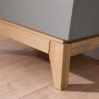 Detailansicht einer PAIDI Babyzimmer Sten – Wickelkommode aus massiver Eiche auf hellem Holzboden, darüber eine graue Verkleidung.