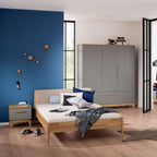Modernes Schlafzimmer mit blauer Akzentwand, Holzbett und PAIDI-Kleiderschrank „Sten“; Kleidung liegt auf dem Boden verstreut.