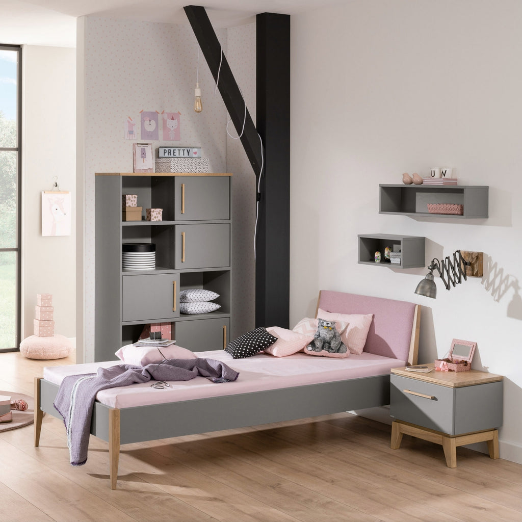 Sonnenlicht beleuchtet ein PAIDI Bettgestell „Sten“ in einem modernen Schlafzimmer in Grau und Rosa mit Regalen aus Massiveiche.
