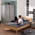 Ein Teenager liest auf dem PAIDI Bettgestell „Sten“ in einem modernen Zimmer; an der Wand sind die Wörter „FRIENDS“ und „LAUGH“ zu sehen.