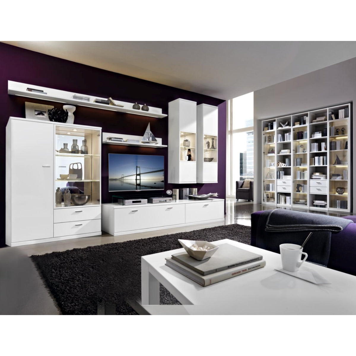 Modernes Wohnzimmer mit der Wohnkombination „RMW Vancouver“, weißen Möbeln, Fernseher, Regalen, Deko, schwarzem Teppich und lila Wand.