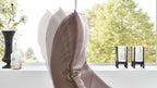 Ein gepolsterter Sessel mit geschwungenen Linien steht vor einem Fenster, daneben zwei Kerzenhalter und eine schwarze Vase mit Blumen.