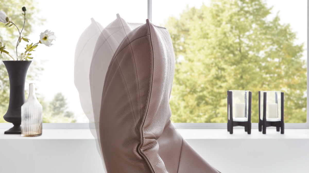 Ein gepolsterter Sessel mit geschwungenen Linien steht vor einem Fenster, daneben zwei Kerzenhalter und eine schwarze Vase mit Blumen.