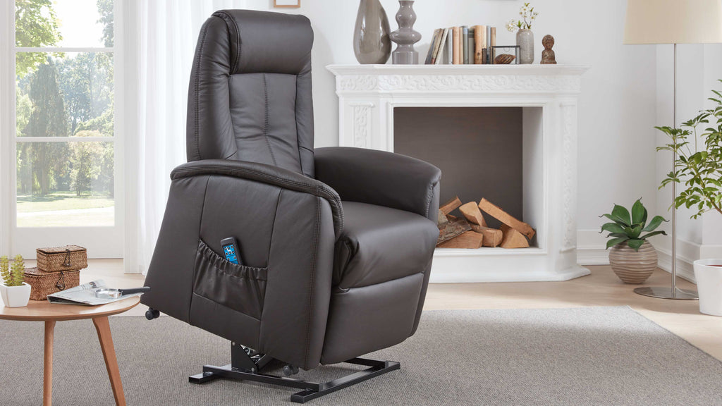 Ein schwarzer Recliner-Stuhl mit armseitiger Fernbedienung steht auf einem grauen Teppich vor einem weißen Kamin, daneben ein runder Tisch mit Pflanzen und einem Zeitschriftenstapel.