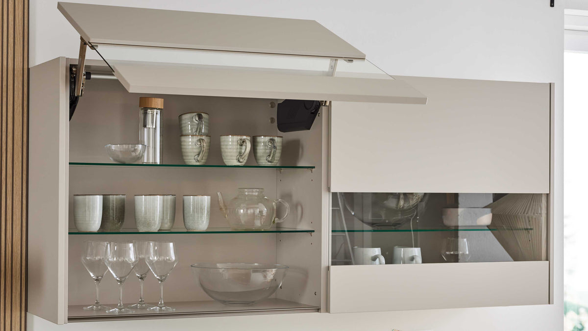 Ein hängender Küchenschrank in beige mit einem Glaseinsatz, gefüllt mit Tassen, Gläsern, einer Karaffe und einer Schüssel.