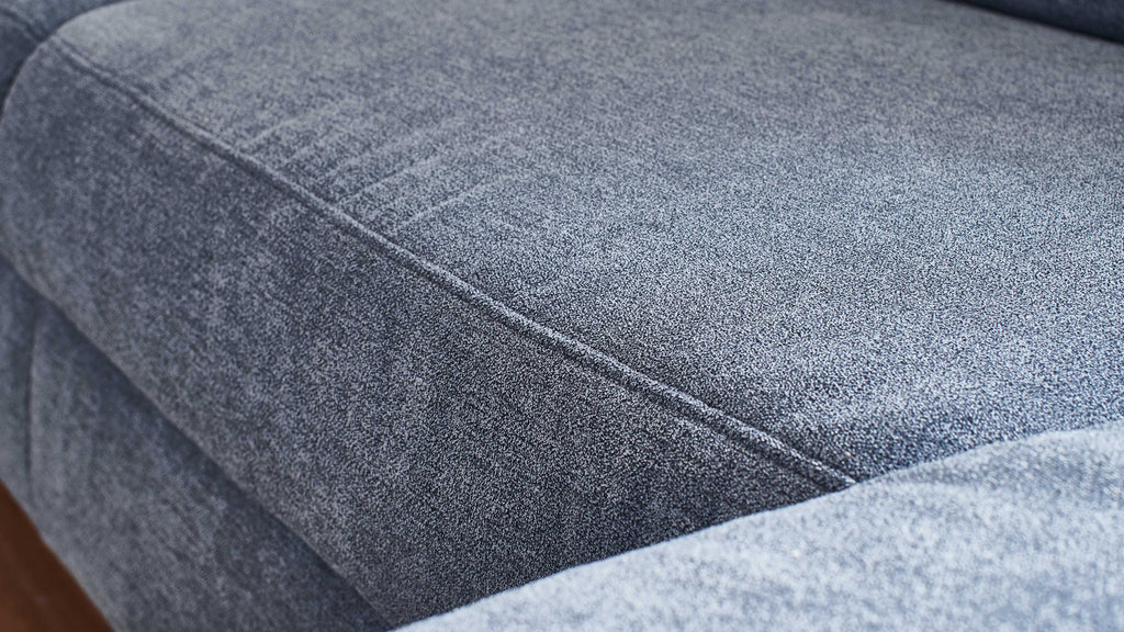Eine graue Couch mit leicht strukturiertem Stoff und sichtbaren Nähten, die auf einer Holzoberfläche steht.