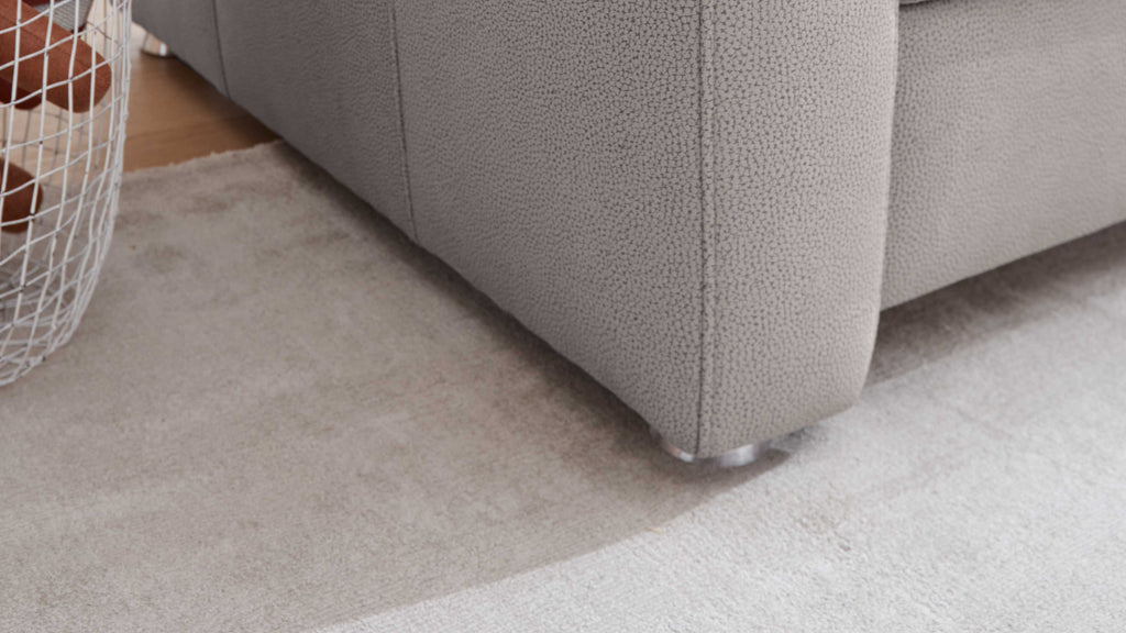 Die Ecke eines grauen Sofas mit strukturiertem Bezug und silbernen Füßen steht auf einem hellen Teppich, daneben ein Drahtkorb mit braunen Kissen.