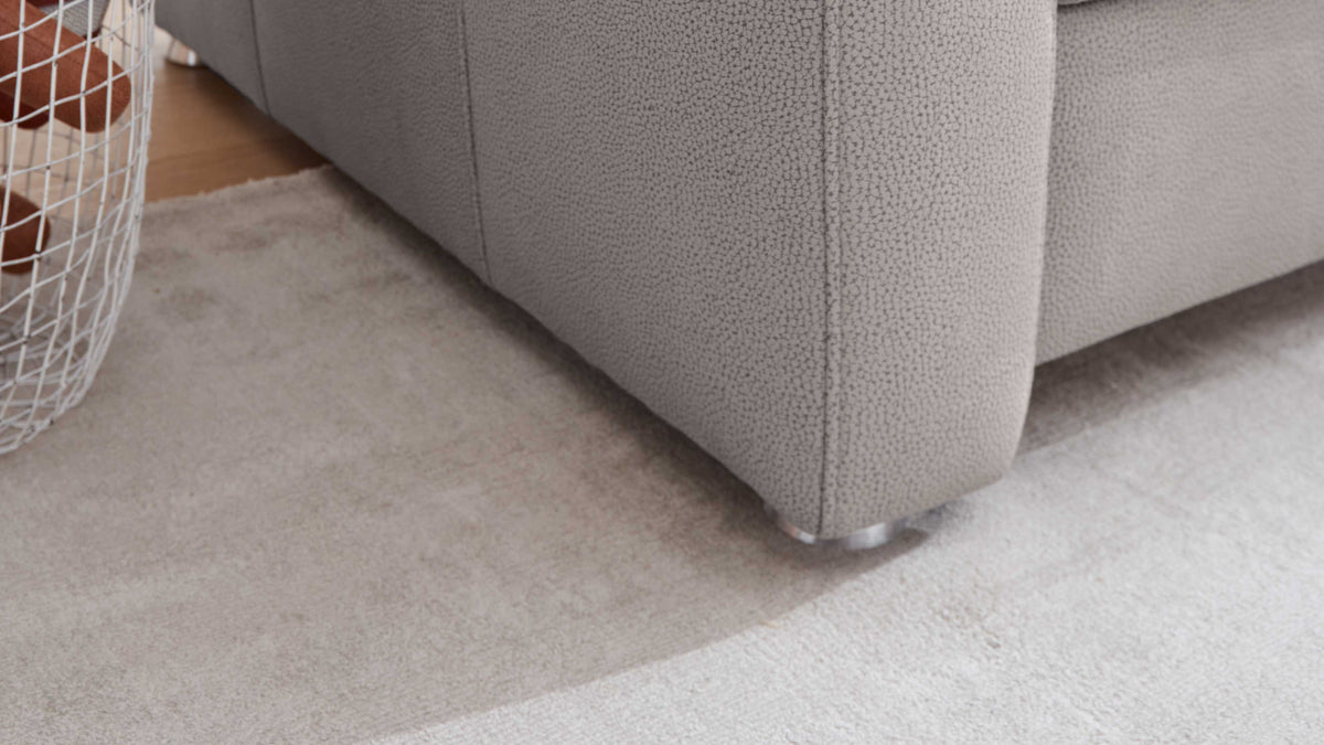 Die Ecke eines grauen Sofas mit strukturiertem Bezug und silbernen Füßen steht auf einem hellen Teppich, daneben ein Drahtkorb mit braunen Kissen.