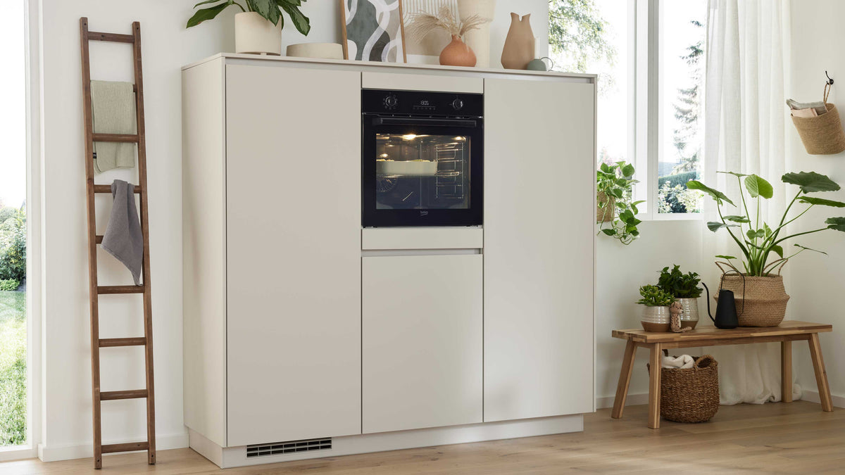 Ein heller Küchenschrank mit integriertem Backofen, daneben eine Holzleiter mit Handtüchern und eine Ablage mit Pflanzen und Körben.