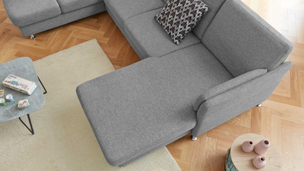 Beige Teppichboden unter einer grauen, L-förmigen Couch mit einem gemusterten Kissen, neben einem Couchtisch mit Dekoartikeln.