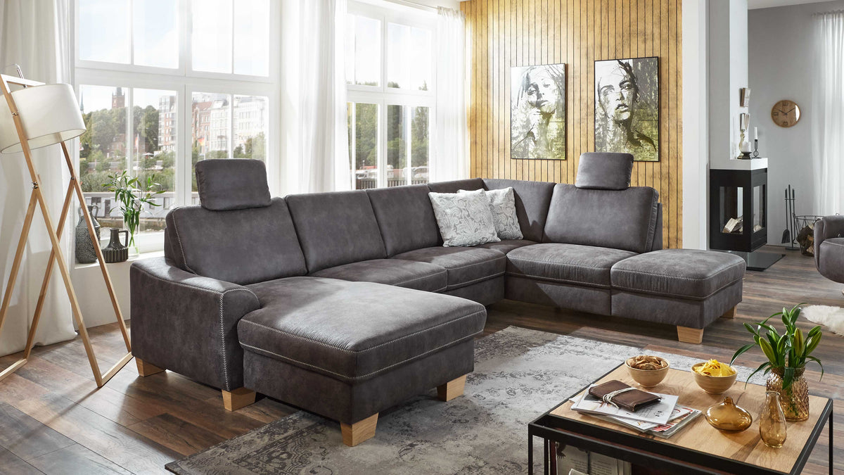 Eine graue L-förmige Couch mit zwei verstellbaren Kopfstützen und Holzbeinen, dekoriert mit einem gemusterten Kissen, steht in einem hellen Raum.