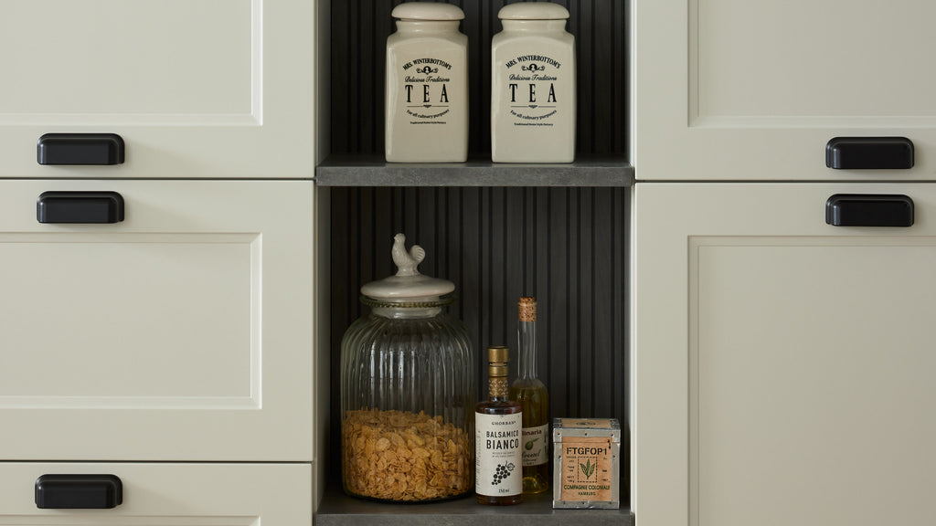 In einem Regal sind zwei Teedosen, ein Glasgefäß mit Haferflocken, eine Flasche Balsamico, eine Ölflasche und eine kleine Schachtel sichtbar.