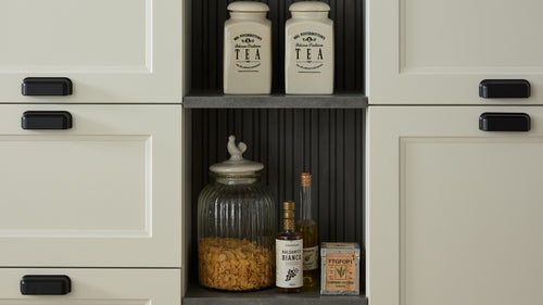 In einem Regal sind zwei Teedosen, ein Glasgefäß mit Haferflocken, eine Flasche Balsamico, eine Ölflasche und eine kleine Schachtel sichtbar.