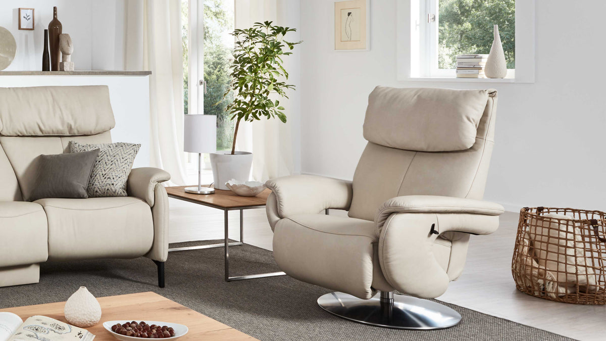 Beige Relaxsessel mit verstellbarem Kopfteil und metallischem Fußgestell, neben einem Holztisch, einer Lampe und einer Zimmerpflanze.