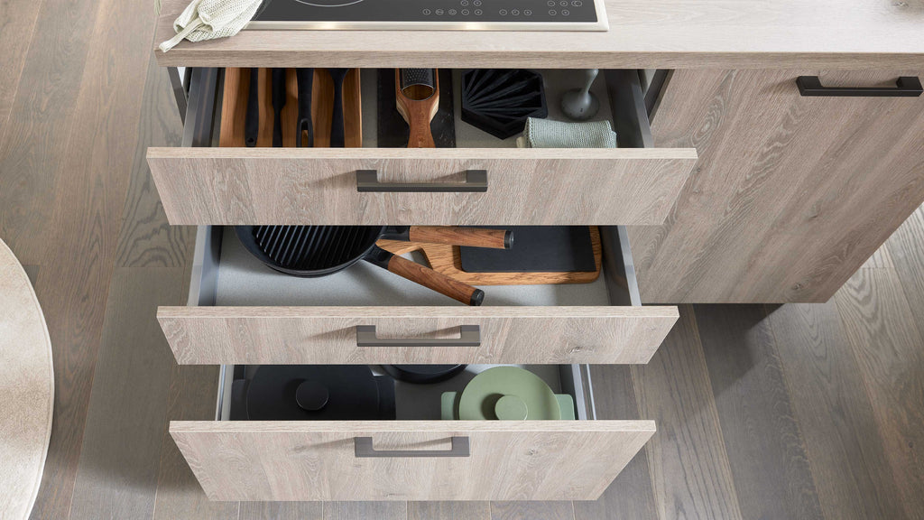 Drei Schubladen einer modernen Küchenzeile sind geöffnet und zeigen Küchenutensilien, Töpfe und einen Kochlöffel aus Holz.
