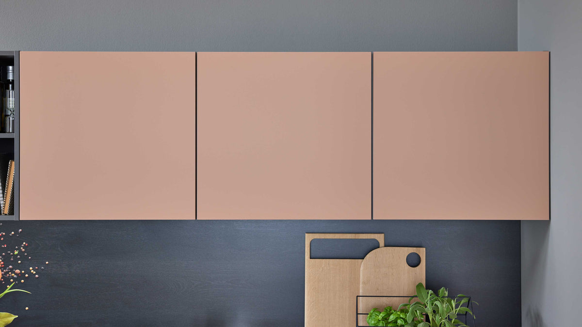 Eine Wand mit drei rosafarbenen Oberschränken und einem schwarzen Küchenunterschrank, daneben Holztabletts und einer kleinen Pflanze.
