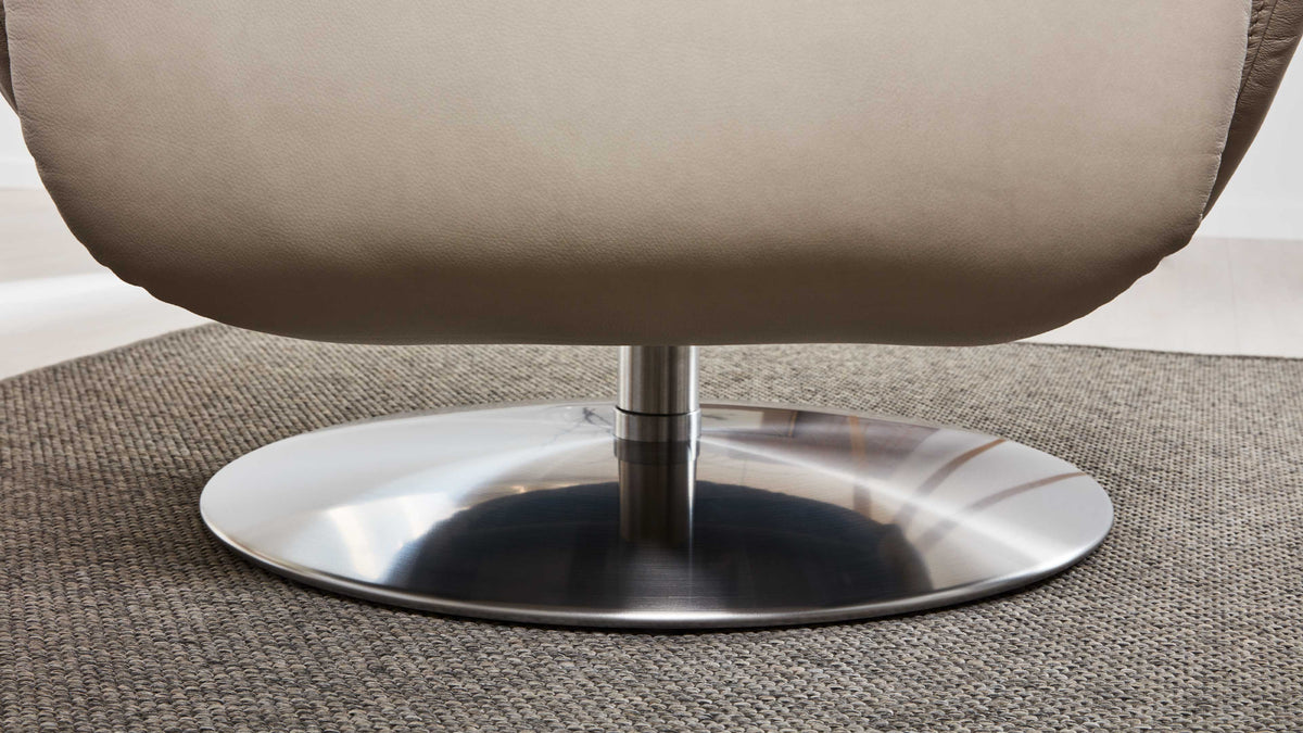 Ein brauner Sessel mit einer runden, silbernen Metallbasis steht auf einem grauen, gewebten Teppich.