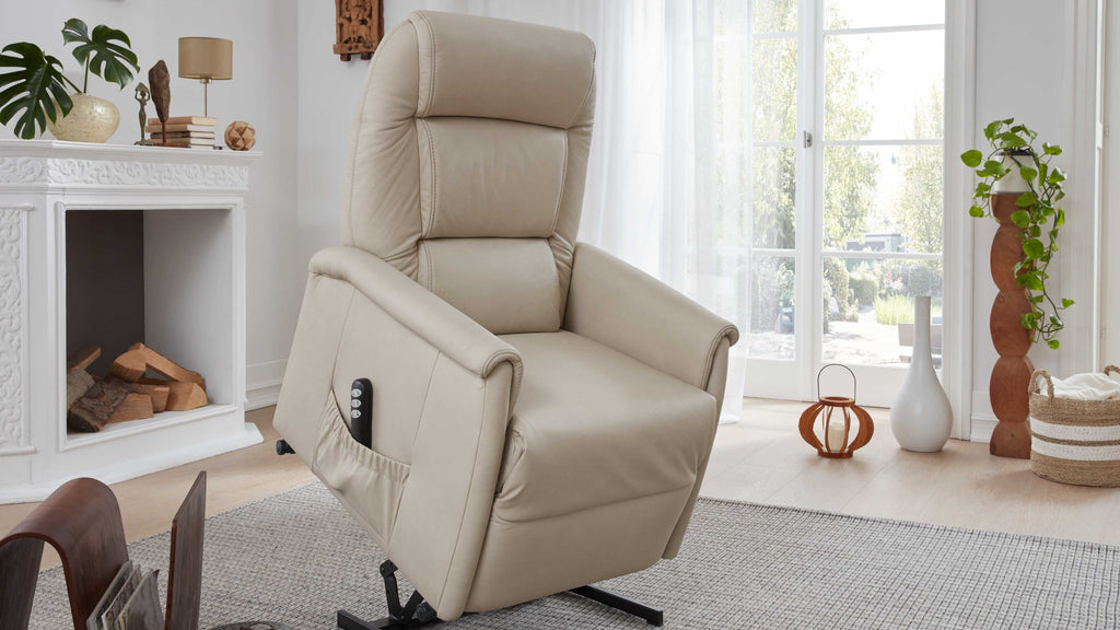 Ein beiger Relaxsessel mit gepolsterter Rückenlehne und seitlichem Bedienelement steht in einem hellen Raum neben einem weißen Beistelltisch und Pflanzen.